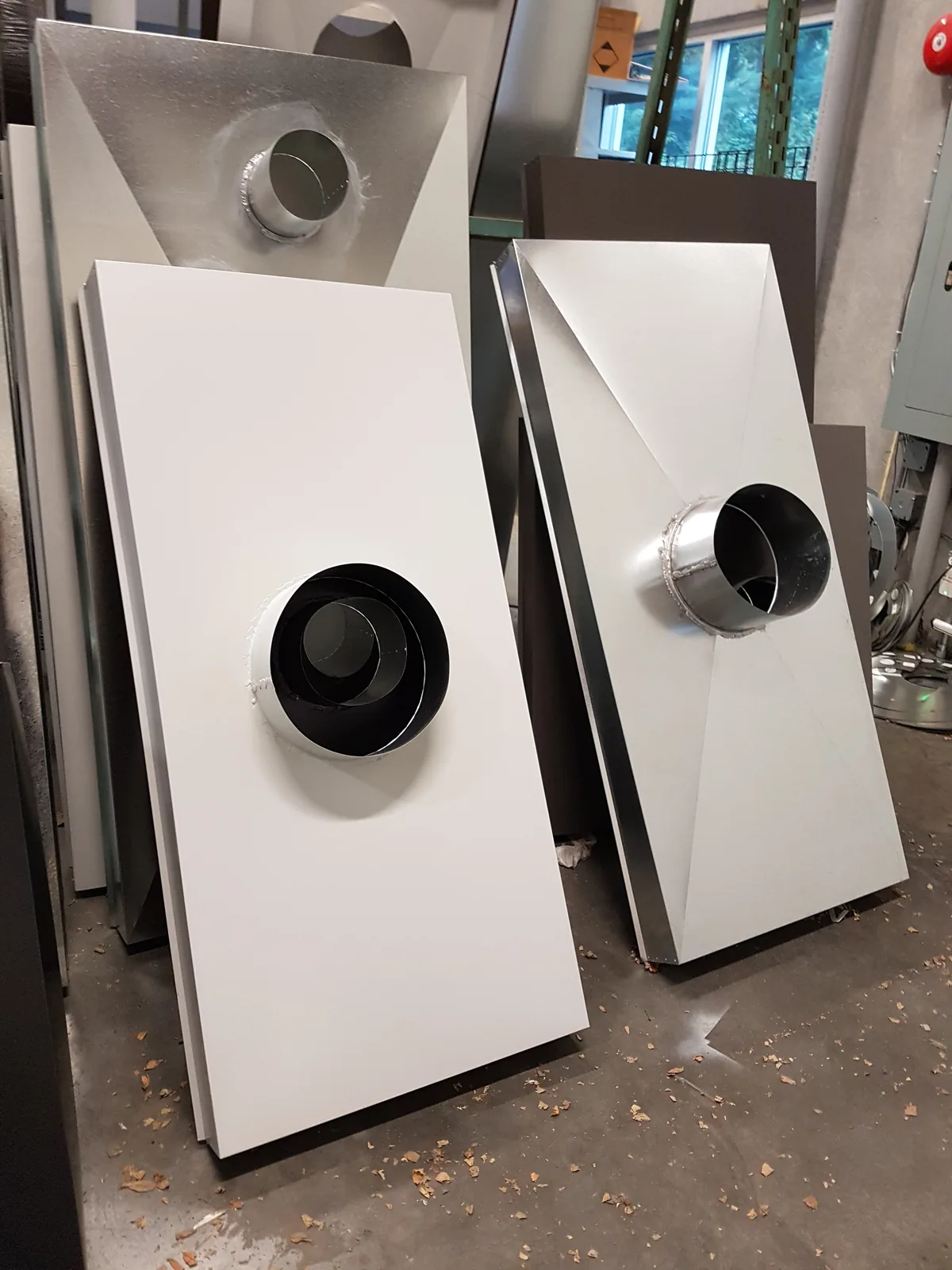 White chimney chase covers standing on the shop floor, each with a sloped hip formed to shed water and a welded round collar for the flue.