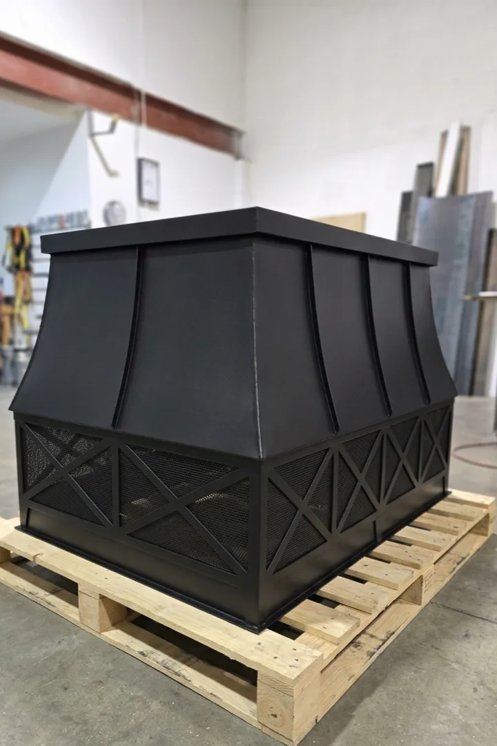 A black architectural chimney shroud on a pallet in the shop, with a concave flared crown, raised seams and a banded base of X-pattern frames over mesh screening.